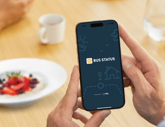 Close up of hands holding a smartphone with the &quot;Bus Status&quot; introductory image appearing on the screen. Background of a coffee cup, water glass and bowl of fruit and oats blurred on a wooden table, with another set of hands showing.