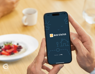 Close up of hands holding a smartphone with the "Bus Status" introductory image appearing on the screen. Background of a coffee cup, water glass and bowl of fruit and oats blurred on a wooden table, with another set of hands showing.