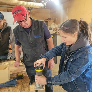 An adult male instructs a female student in woodworking as she uses a power tool in a wood shop at the Consul School's student convention day