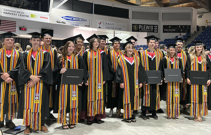 Fifteen Grade 12 students pose together at a graduation ceremony, wearing their caps and gowns decorated with a Fransaskois arrow belt. Some students are holding their diplomas.