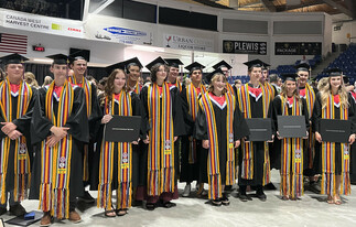 Fifteen Grade 12 students pose together at a graduation ceremony, wearing their caps and gowns decorated with a Fransaskois arrow belt. Some students are holding their diplomas.