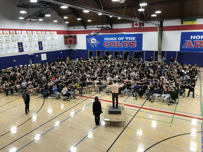 Band conductor on a platform raising his baton and directs 560 band and choral students of various ages in front of him to play and sing. Two other band directors are standing off to the left.